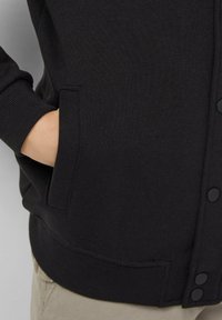 Czarny kardigan o fakturowanym splocie, z zapięciem na guziki z przodu oraz kieszeniami po bokach. Mankiety i dół są prążkowane dla pewnego dopasowania.