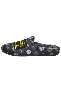 Pantufla de tela negra con interior de felpa, con patrones de calaveras en gris y blanco, y un gráfico de texto amarillo en la parte superior. Suela plana.