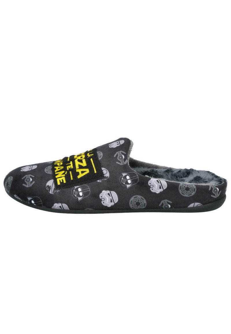 Pantufla de tela negra con interior de felpa, con patrones de calaveras en gris y blanco, y un gráfico de texto amarillo en la parte superior. Suela plana.