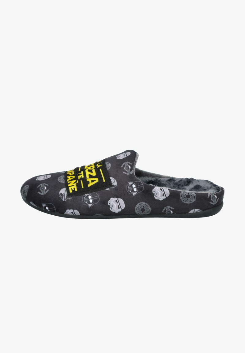 Pantufla de tela negra con interior de felpa, con patrones de calaveras en gris y blanco, y un gráfico de texto amarillo en la parte superior. Suela plana.