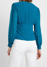 Blouse teal à manches longues avec des épaules bouffantes et un détail froncé au dos. Le tissu semble lisse avec un léger éclat. Associée à un jean blanc.