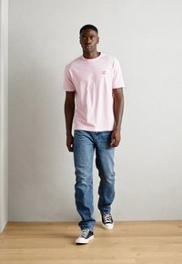 Roze katoenen t-shirt met een klein hartlogo, gecombineerd met blauwe spijkerbroek en zwarte sneakers. Model staat tegen een lichtgrijze muur.