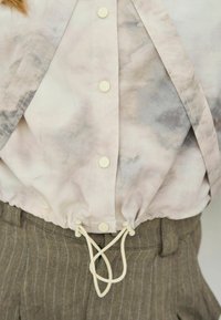 Camicia color crema con leggero motivo, bottoni e coulisse regolabile all'orlo, abbinata a pantaloni marroni a righe sottili.