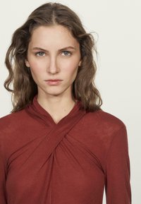 Červený top s dlouhým rukávem a zavinovacím designem, s texturovanou tkaninou a vysokým výstřihem se nařasenými detaily na hrudi.