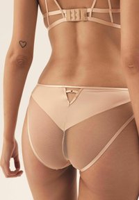 Rücken einer Frau in hautfarbener, transparenter Mesh-Bikiniunterwäsche und passendem BH mit einem Herz-Tattoo am linken Arm.