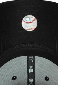 Schwarze Baseballmütze mit grauer Unterseite. Im Inneren befindet sich ein runder MLB-Aufkleber, der einem Baseball ähnelt. New Era-Branding ist ebenfalls vorhanden.