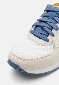 Sport sneaker featuring a mix of white mesh and beige suede with blue laces, a light-colored midsole, and a textured blue rubber outsole.