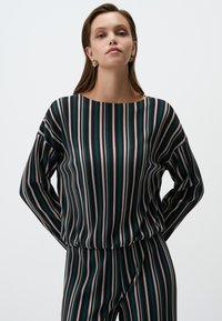 Blusa a righe in verde scuro, beige e turchese. Presenta maniche lunghe e un ampia scollatura. Tessuto liscio e leggero con righe verticali.