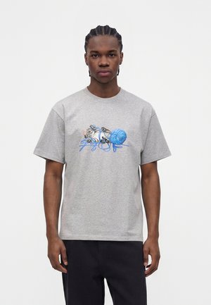 Homme aux cheveux tressés portant un t-shirt gris avec un chaton emmêlé dans du fil bleu, debout devant un fond blanc uni.