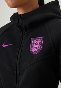 Melnā kapucēta džemperis ar rāvējslēdzēju, kuram ir purpursarkans Nike logo un izsist purpursarkans vairogs ar lauva motīviem. Izgatavots no teksturēta auduma ar šuvju detaļām.