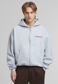 Hoodie gris zippé avec une poche avant, coupe oversize, et "PROHIBÉ" en lettres rouges sur la poitrine. Tissu doux, design décontracté.