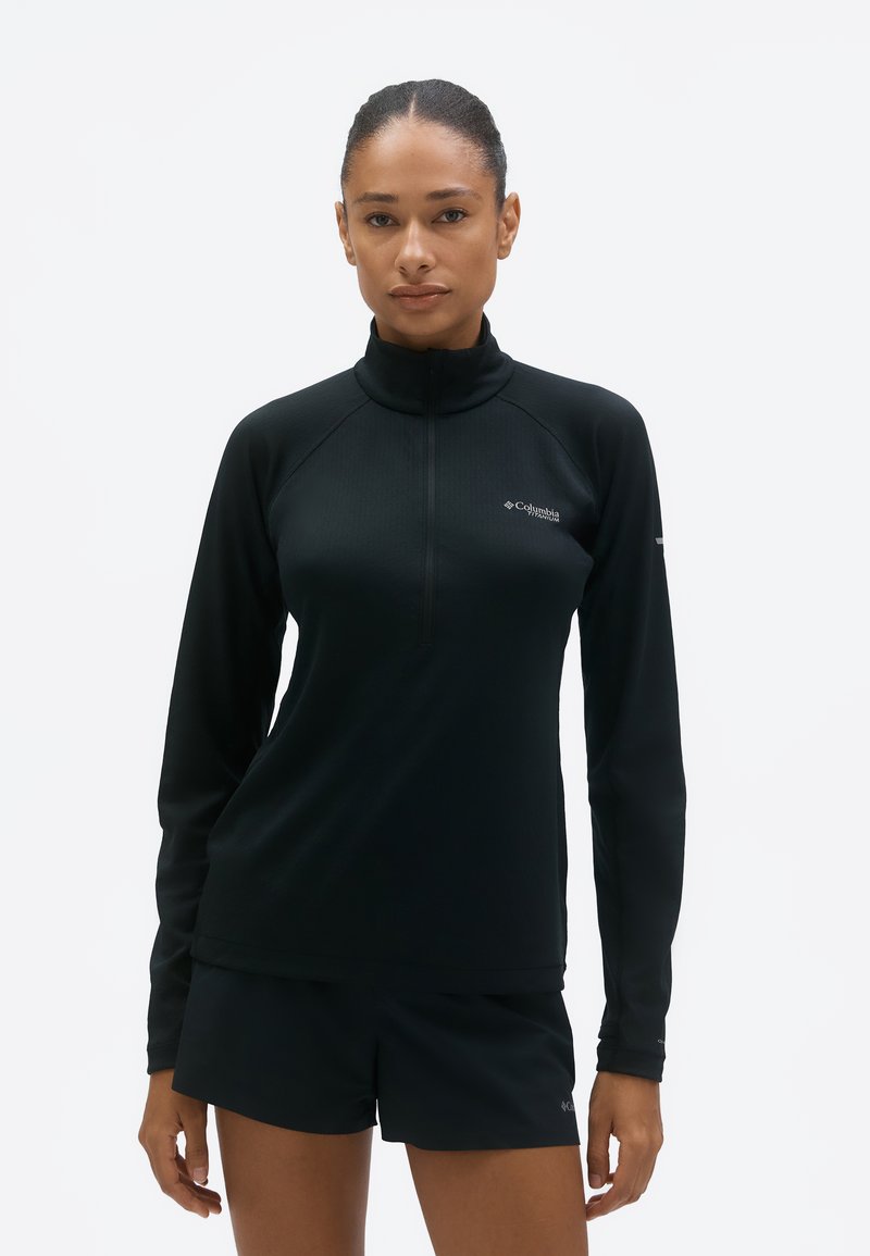 Woman standing facing forward wearing a black long-sleeve zip-up top and black shorts against a plain white background.