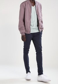 Light mauve bomber jacket, light green V-neck t-shirt, navy slim-fit pants, and white sneakers. Smooth fabric and streamlined design.