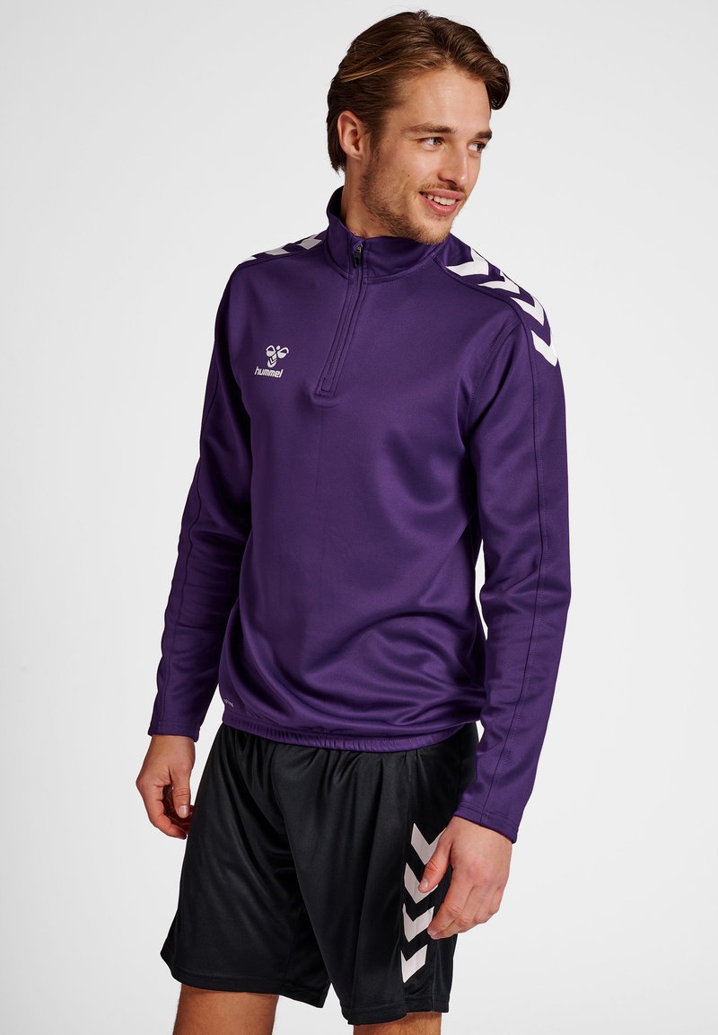 Homme portant un haut de sport violet à fermeture éclair quart, avec des bandes chevrons blanches sur les épaules et un short noir avec motif chevron blanc.