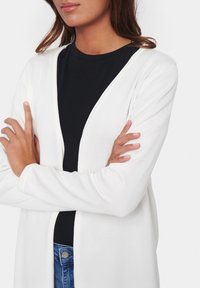 Kvinna med medelhudton iförd vit cardigan över svart skjorta och blå jeans, med armarna korsade över bröstet.