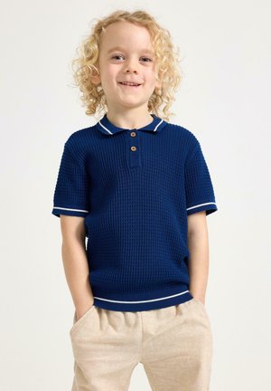 Smilende barn med krøllet blondt hår iført en marineblå kortærmet strikket polo og beige bukser, hænderne i lommerne, stående mod en ensfarvet baggrund.