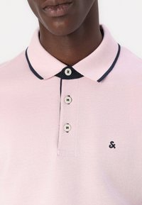 Roze polo shirt van een textuurstof, met een marineblauwe kraag en drie knopen. Een geborduurd logo is geplaatst op de linkerkant van de borst.