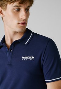 Marineblauw polo shirt met een kraag met twee witte strepen. Kenmerkt het Red Bull Racing logo in het wit, gemaakt van een gestructureerde, ademende stof.