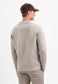 Beige pullover, zachte stof, lange mouwen, achterkant. Bruine pet met een gestructureerde afwerking, licht gebogen rand, verstelbare strap aan de achterkant.