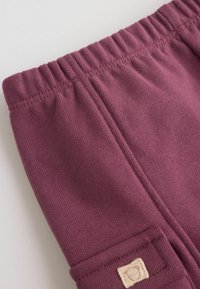 Gros plan d’un pantalon à taille élastique bordeaux avec une couture verticale visible et un petit patch beige avec un logo de visage d’animal.