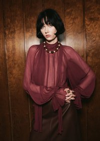 Mlada ženska s kratkimi črnimi lasmi nosi prosojno bordo blouse z obsežnimi rokavi, temen krilo in ogrlico iz kroglic, stoji ob leseni steni.