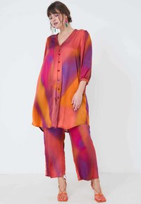 Vloeiende outfit in levendige oranje en paarse tinten. Satijnen stof, losse pasvorm, lange mouwen met knopen, gecombineerd met bijpassende wijde broek en sandalen.