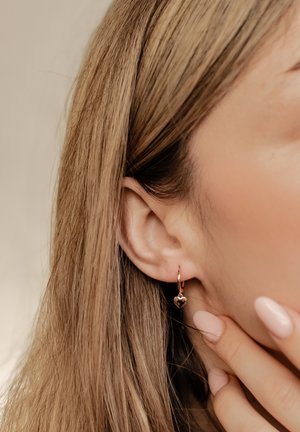 Orecchini a cerchio in oro rosa con un piccolo ciondolo a forma di cuore, indossati da una persona con lunghi capelli castano chiaro e smalto per unghie neutro.