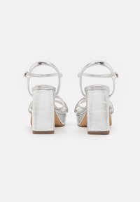Silver metallic block-heeled sandals with ankle straps, featuring a smooth texture and circular accents on the straps.