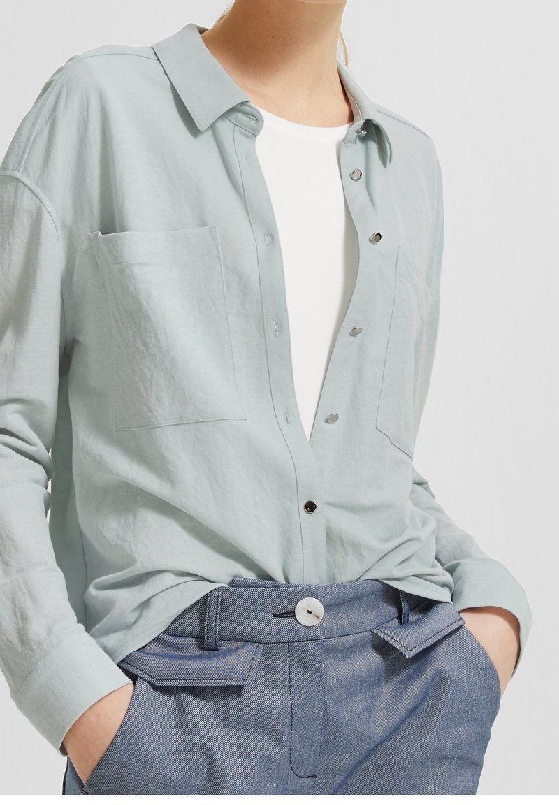 Personne portant une chemise bleu clair à boutons par-dessus un haut blanc, rentrée dans un pantalon bleu taille haute, les mains dans les poches.