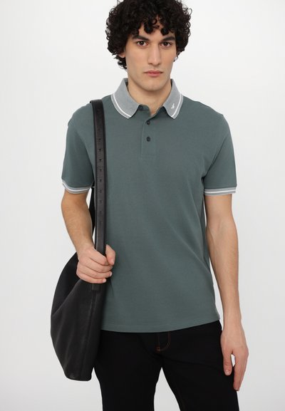 Grønn polo skjorte med stripete krage, kort ermer og tre-knapps placket. Modellen holder en svart lærveske, stående mot en enkel bakgrunn.