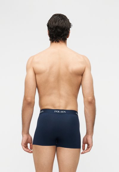 Bărbat cu păr scurt și închis la culoare, cu spatele gol, purtând boxeri bleumarin cu textul „POL ØREN” pe banda din talie, stând în fața unui fundal simplu și deschis la culoare.