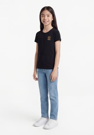 Naeratav tüdruk seisab, kandes musta Versace logoga T-särki, hele siniseid teksapükse ja valgeid tenniseid valgel taustal.