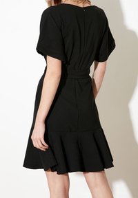 Femme portant une robe noire à manches courtes avec une taille cintrée et un ourlet évasé, se tenant devant un fond blanc avec une ombre minimale.