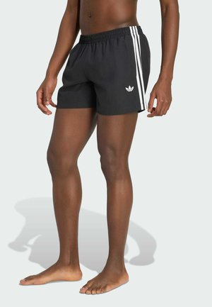 Persona descalza que lleva pantalones cortos negros de Adidas con tres rayas blancas y el logo, de pie frente a un fondo claro sencillo.