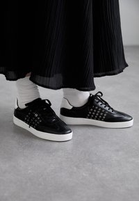 Baskets en cuir noir avec des œillets argentés, semelles en caoutchouc blanc et lacets noirs, associées à des chaussettes blanches et une jupe plissée noire.