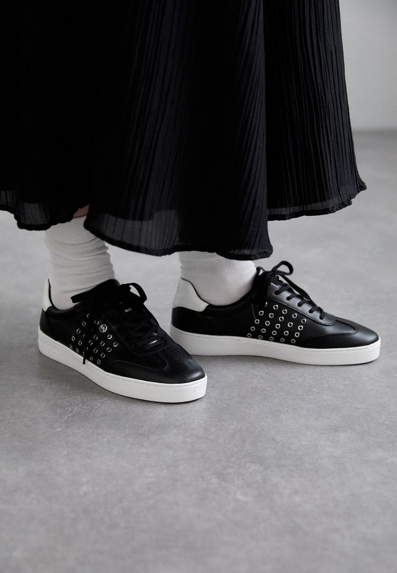 Baskets en cuir noir avec des œillets argentés, semelles en caoutchouc blanc et lacets noirs, associées à des chaussettes blanches et une jupe plissée noire.