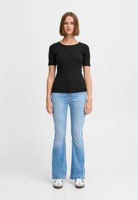 Top corto a maniche corte nero a coste abbinato a jeans flare azzurri e sneakers bianche, indossato da una persona in piedi contro uno sfondo neutro.