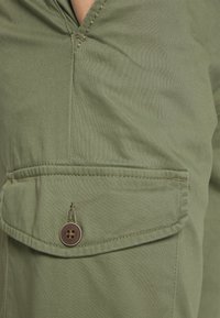 Olive green button-up shirt featuring a chest pocket with a brown button and stitching details, made from a smooth, textured fabric.