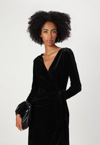 Vestido envolvente preto de veludo com mangas longas, decote em V profundo e detalhe de cintura amarrada. Modelo segura uma clutch preta. Textura suave, design elegante.