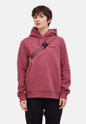 Personne aux cheveux courts portant un sweat à capuche rose poudré et un pantalon noir, portant un sac à bandoulière assorti avec le logo "Peak Performance".