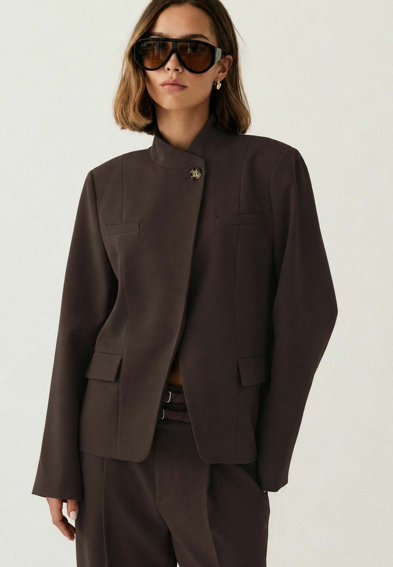Γυναίκα που φοράει υπερμεγέθη σκουρό καφέ σακάκι και ταιριαστό παντελόνι, μεγάλα μαύρα γυαλιά ηλίου και χρυσά κρίκους σκουλαρίκια, στέκεται μπροστά σε απλό φόντο.