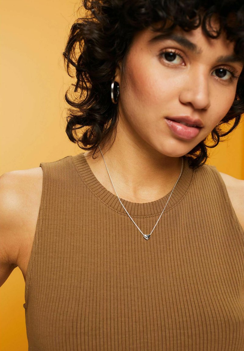Top in cotone a coste marrone, collana d'argento con pendente a forma di cuore, orecchino a cerchio e capelli ricci su uno sfondo giallo.