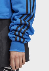 Sweat-shirt bleu avec des poignets côtelés noirs et trois rayures noires le long de la manche. Le matériau semble doux avec une texture lisse.