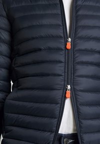 Giubbotto imbottito di colore blu navy con cuciture orizzontali, dotato di una zip frontale con dettagli arancioni e argento. L'interno è bianco.