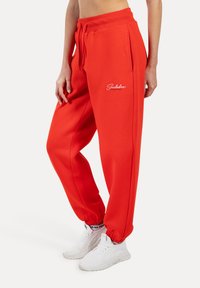 Červené joggingové kalhoty ve volném střihu, s elastickým pasem na šňůrku, bočními kapsami a bílým logem značky na levém stehně.