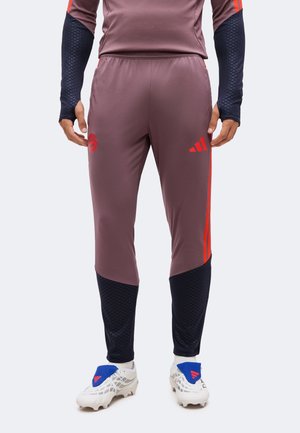 Atleta con pantalones de entrenamiento rojo púrpura con parches negros, tacos blancos con cubiertas azules y una camiseta de manga larga a juego.