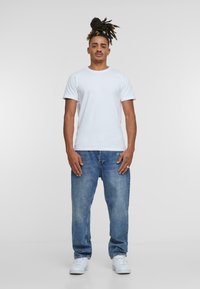 T-shirt bianco a maniche corte abbinato a jeans denim azzurri chiari. I jeans hanno una vestibilità rilassata e un leggero effetto usurato, insieme a sneakers bianche.