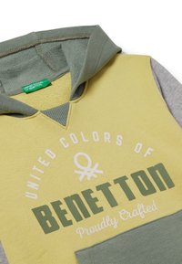 Felpa gialla e verde con cappuccio, con il testo "BENETTON" in verde scuro. Tessuto morbido e maniche grigie. Etichetta visibile all'interno.