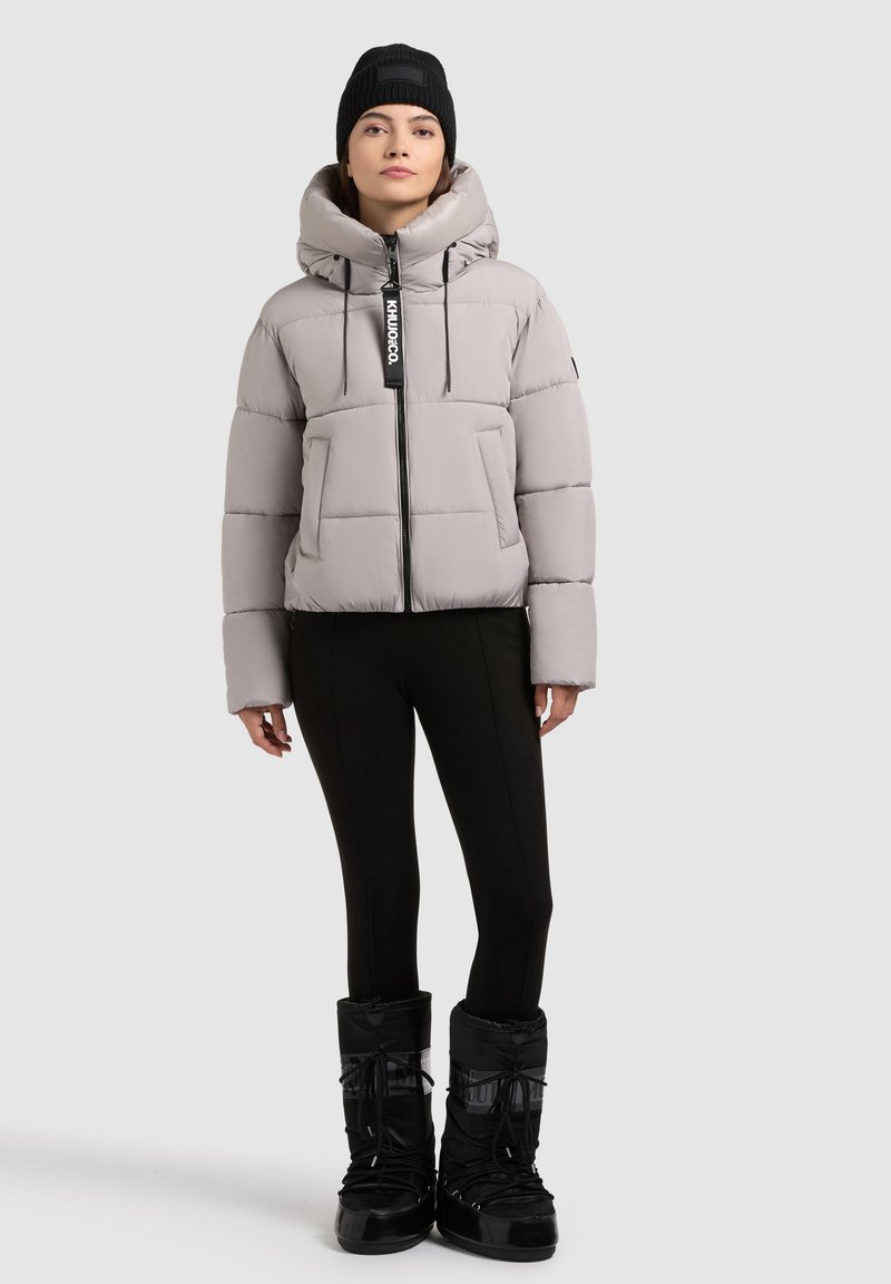 Giacca a vento leggera grigia chiara con colletto alto, tasche frontali e zip. Leggings neri e stivali da neve neri abbinati con lacci.