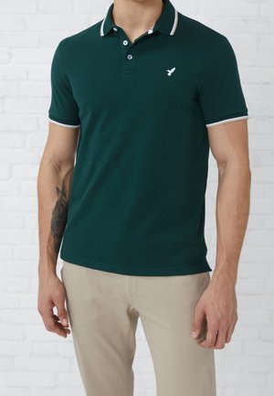 Mann trägt dunkelgrünes Polohemd mit kurzen Ärmeln und weißen Kanten sowie beige Hosen, steht vor einer weißen Ziegelmauer.